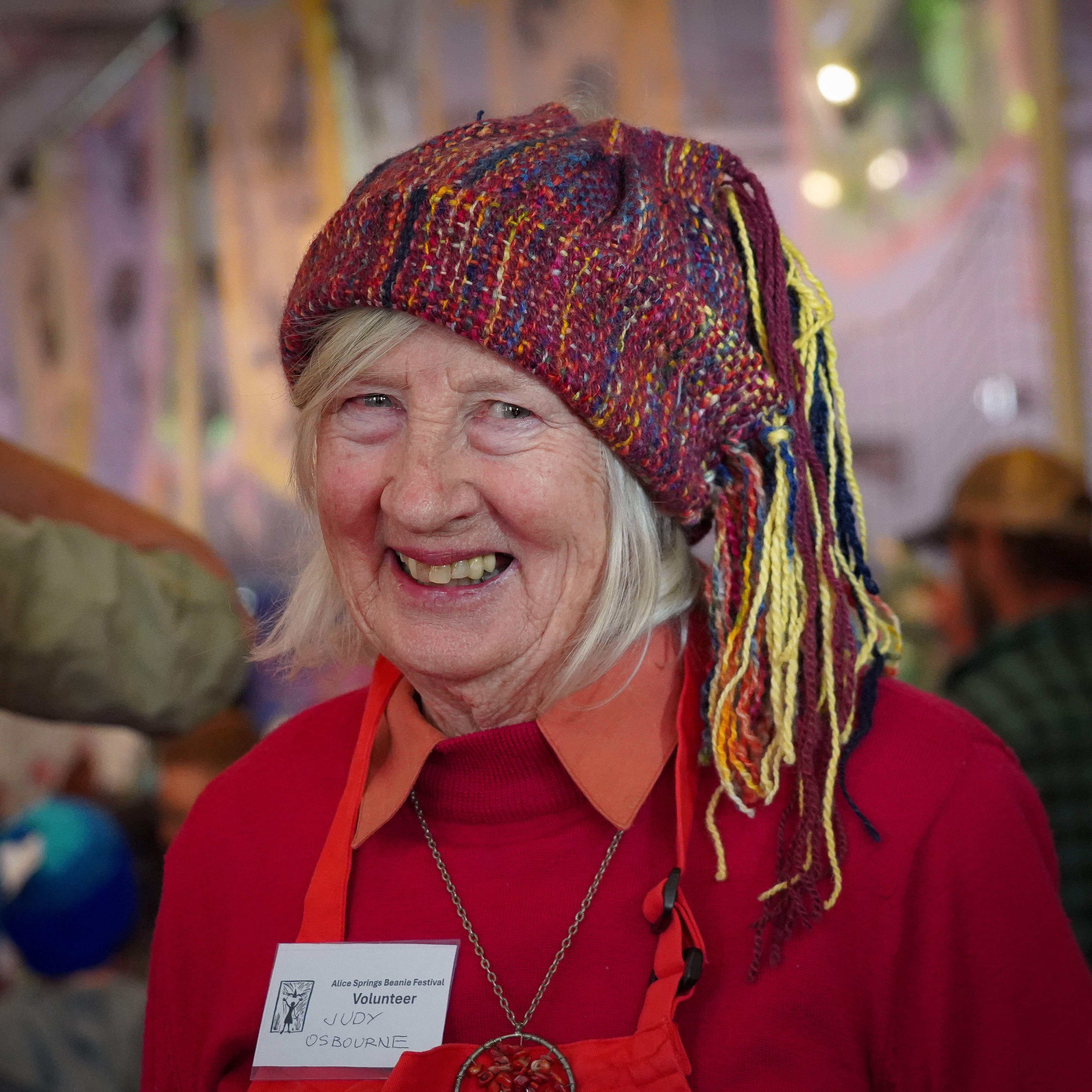 A woman smiles at the camera while wearing a colourful knitted beanie, strands of yarn are sewn on top of the hat.
