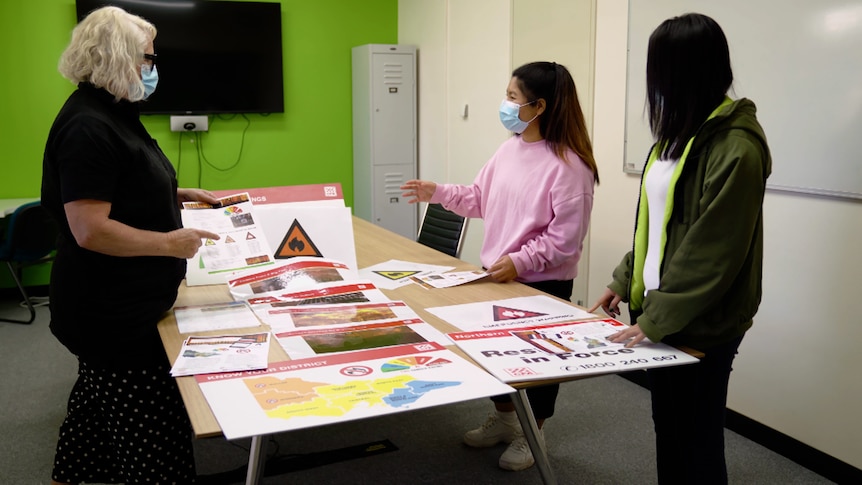 Ku Htee is seen wearing a pink jumper and blue mask and gesturing as she stands at a table with two other women.
