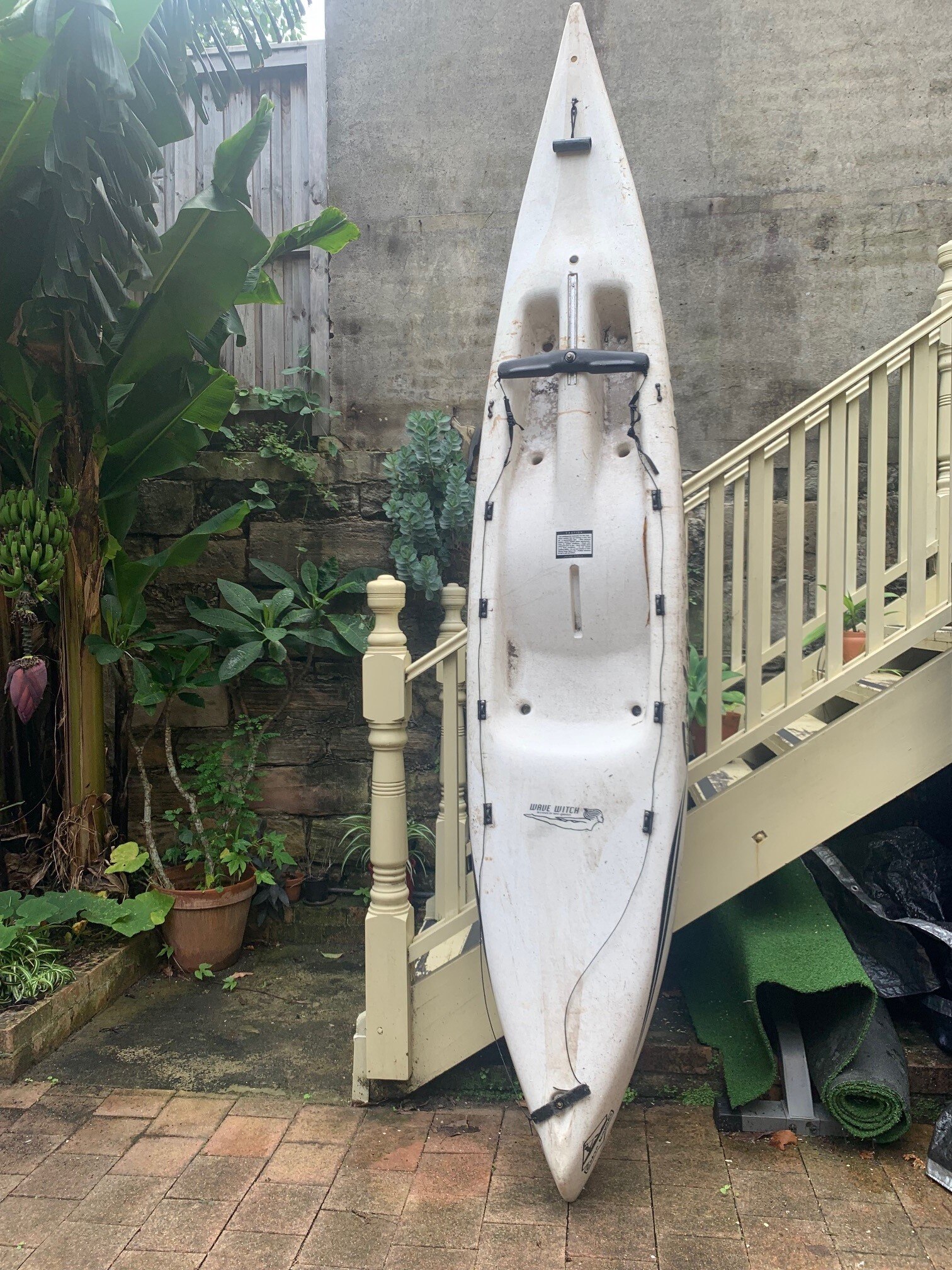 A white Wave Witch Kayak leaning on pale yellow stairs in a backyard.