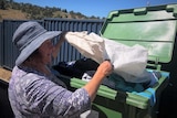 A woman in a blue hat tips clothes out of a white polyester bag into a large green bin.
