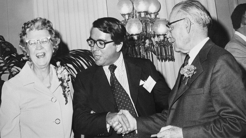 Black and white photo of man wearing suit and glasses, Robert Bauman, standing next to older man and woman. 