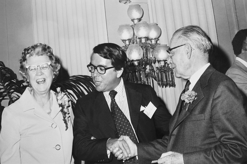 Black and white photo of man wearing suit and glasses, Robert Bauman, standing next to older man and woman. 