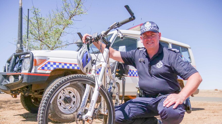 A police officer kneels next to a police bicycle, in front of his car in the rural Queensland.