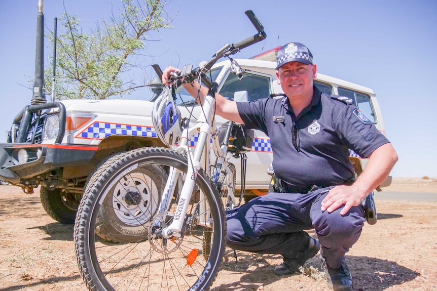 A police officer kneels next to a police bicycle, in front of his car in the rural Queensland.