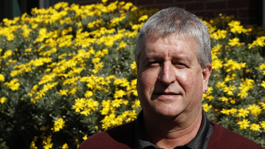 a man with grey hair wearing a burgundy jumper stands in front of a bush of yellow flowers