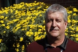 a man with grey hair wearing a burgundy jumper stands in front of a bush of yellow flowers