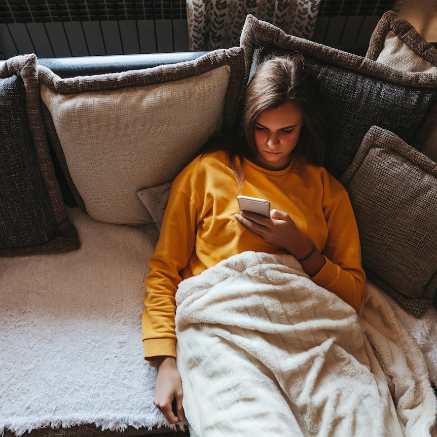 Woman in a yellow jumper sits on a grey couch looking at her phone. 