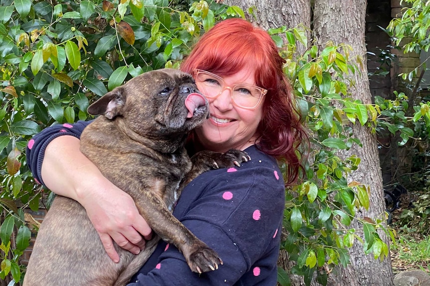 A woman with copper-coloured hair and spectacles hugs a dog while standing in front of a tree.