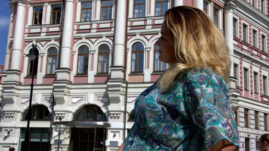 A blonde woman in a white and blue paisley shirt looks into the distance in front of russian architecture in moscow.