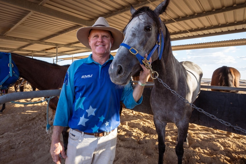A smiling man in a blue shirt, white jeans, Akubra hat stands with horse in a stable with other horses around him.