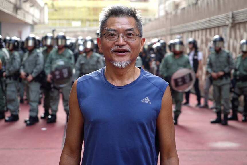 William Cheung smiles as he stands in front of riot police wearing a blue Adidas singlet. He has grey hair and wears glasses.