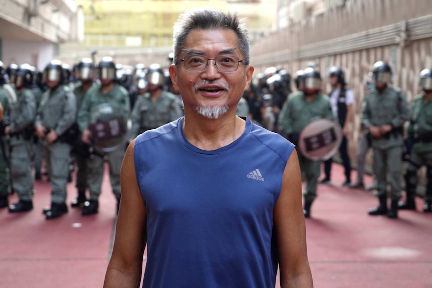William Cheung smiles as he stands in front of riot police wearing a blue Adidas singlet. He has grey hair and wears glasses.