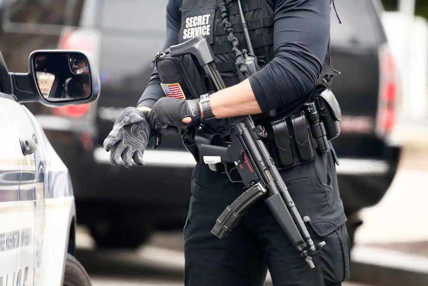 View of the torso and top of the legs of a secret service officer in uniform with a large gun, pulling on a glove.