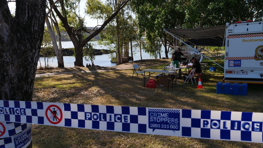 Police tape in front of a river bank with emergency services set up with tables
