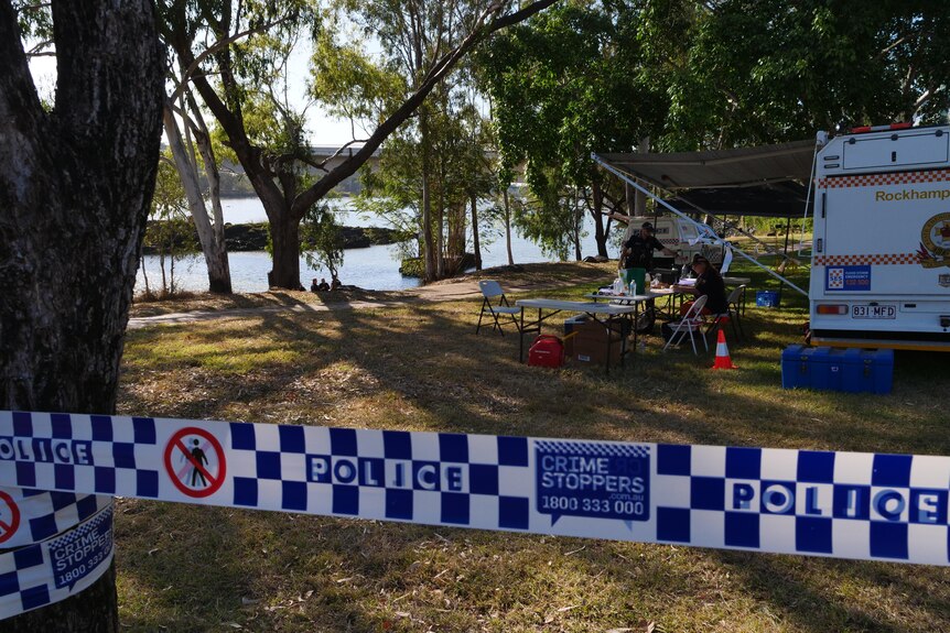 Police tape in front of a river bank with emergency services set up with tables