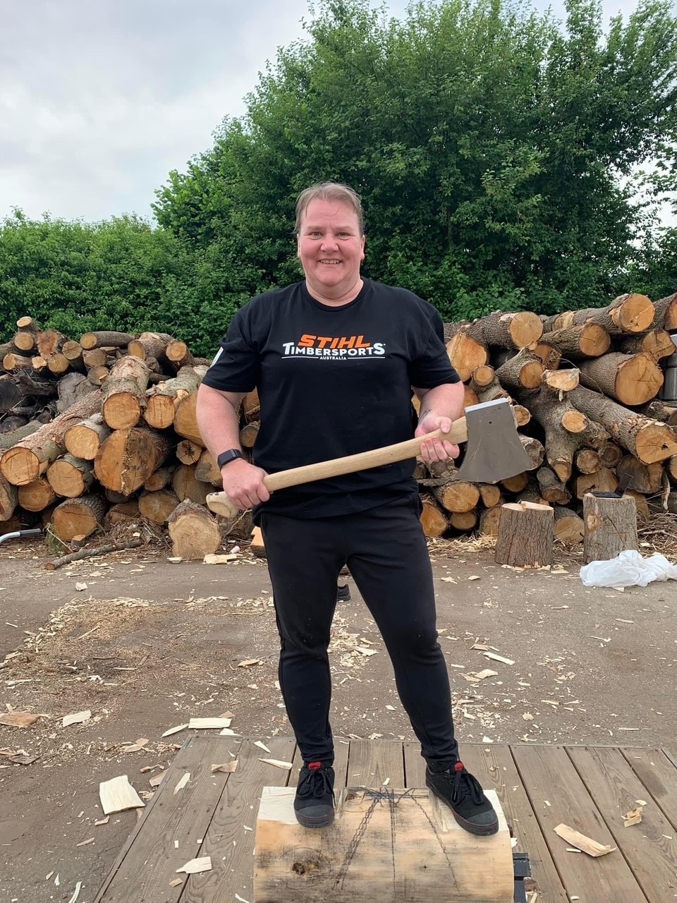 mulher está segurando um machado vestindo uma camiseta preta e calça e sorri para a câmera 