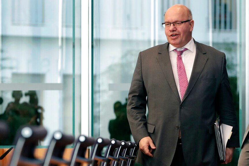 German Economy Minister Peter Altmaier walks past a row of empty boardroom chairs in a grey suit and loose pink tie.