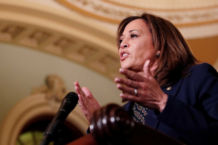 Kamala Harris stands at a lectern, gesturing with her hands and talking to reporters.