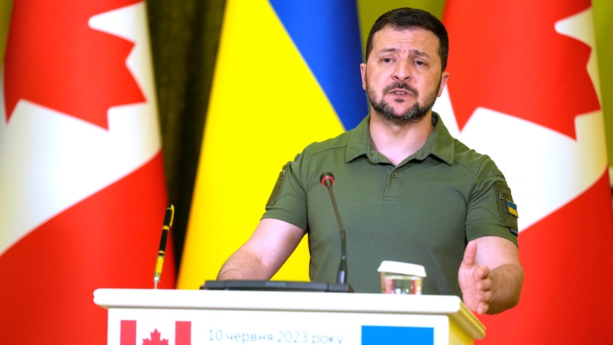 A middle-aged Ukrainian man in a tight combat polo shirt stands behind a podium in front of Ukrainian and Canadian flags.