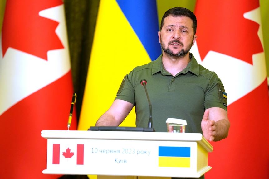 A middle-aged Ukrainian man in a tight combat polo shirt stands behind a podium in front of Ukrainian and Canadian flags.
