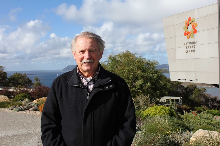 A man stands in front of the National ANZAC Centre, with the ocean in the background
