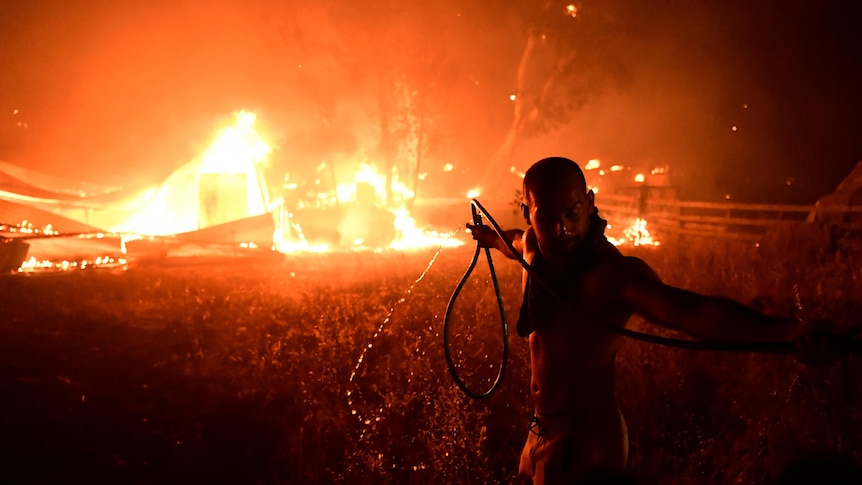 The silhouette of a man pulling a hose is backlit by a structure on fire behind him