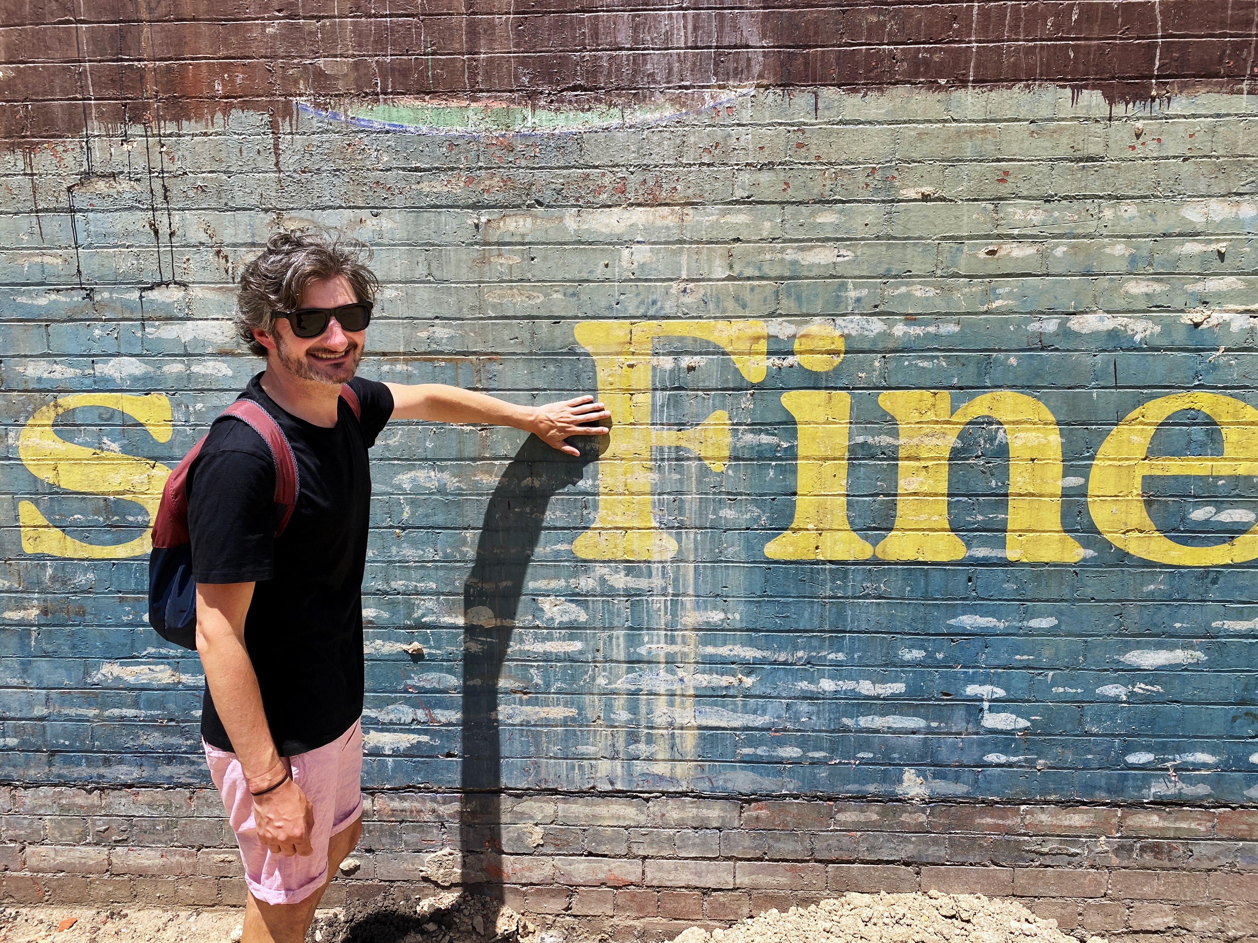 A smiling man pointing at an old blue and yellow sign.