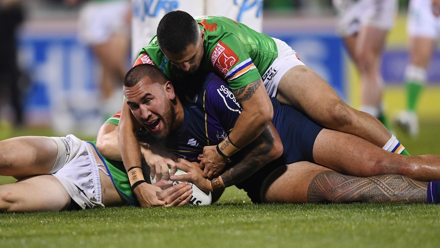 A Melbourne Storm NRL player lies on the ground after scoring a try with two Canberra opponents on top of him.