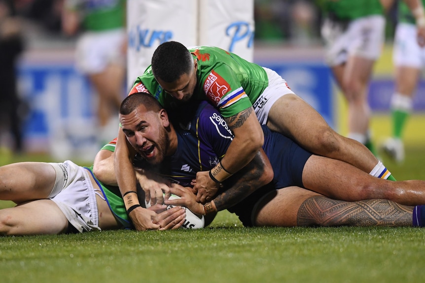 A Melbourne Storm NRL player lies on the ground after scoring a try with two Canberra opponents on top of him.