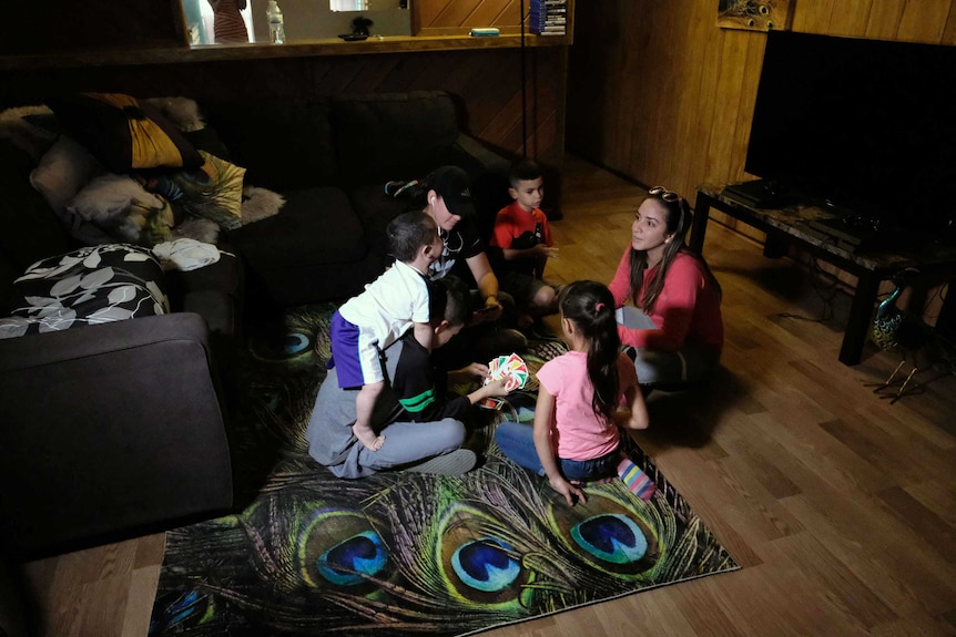 A group of young people sit inside a small, dark room. It is filled with furniture, including a couch, tv and a bright rug.