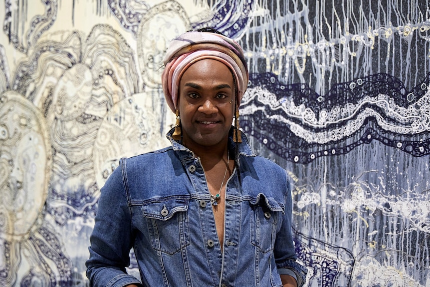 An Indigenous Australian man in a denim jacket and colourful head wrap smiles in front of a painting.