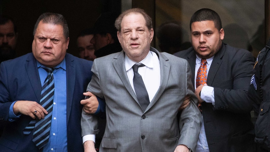 Harvey Weinstein wears a suit and is supported by two man holding him up via each arm