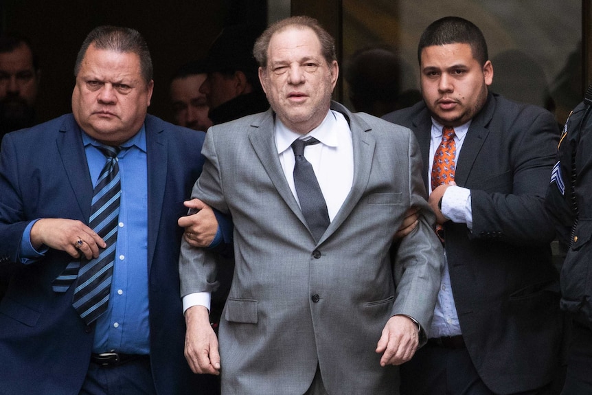 Harvey Weinstein wears a suit and is supported by two man holding him up via each arm