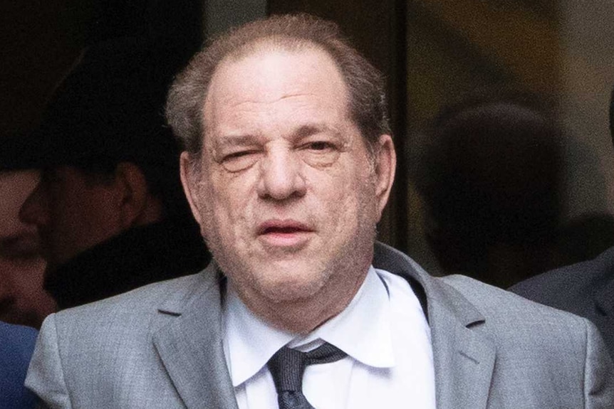 Harvey Weinstein wears a suit and is supported by two man holding him up via each arm