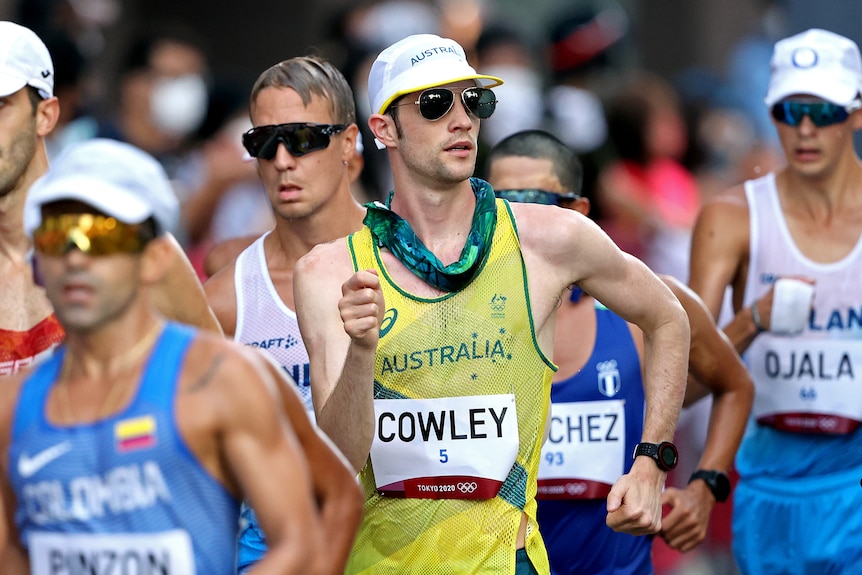 Rhydian Cowley, wearing a hat and sunglasses, and drenched in water, in the middle of a pack of race walkers.
