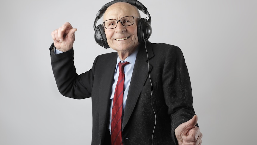 Older man wearing a suit and tie, he has a bald head and glasses and is dancing with headphones on.