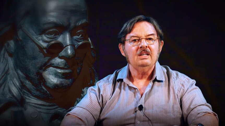 Man in glasses and striped shirt speaks to the camera with an image of a statue in the background.