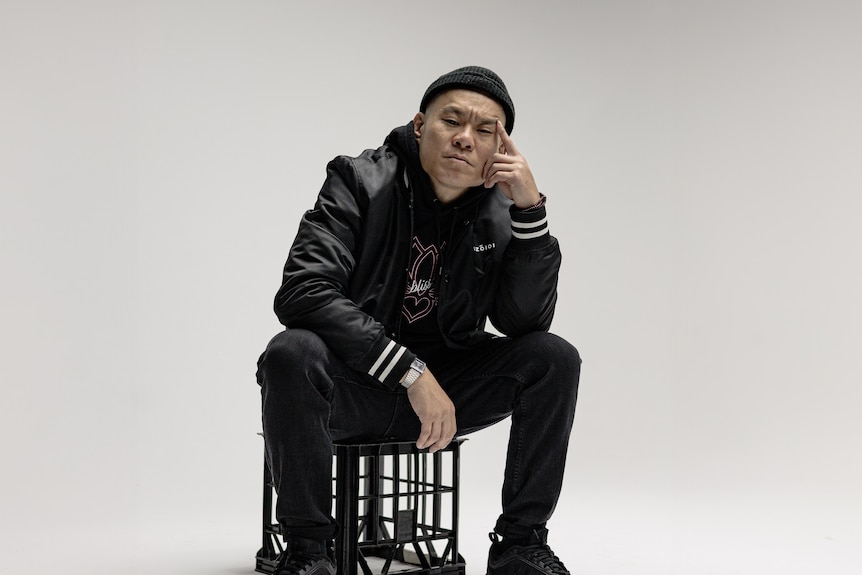 An asian man on a milk crate, wears all black, cap, jacket with white stripes on sleeve.Thinking pose, seroius.
