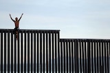 Man sits on top of the US-Mexico border fence with his hands in the air