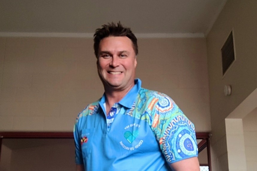 A man smiles. He wears a light blue polo shirt with an Indigenous design on the sleeve.