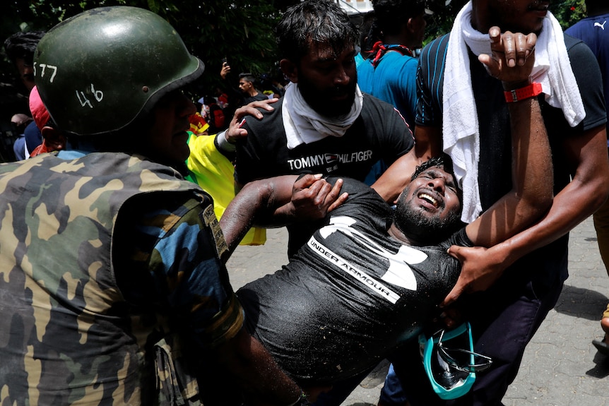 Three people, one wearing apparent military clothing, carry a man wincing in pain. 