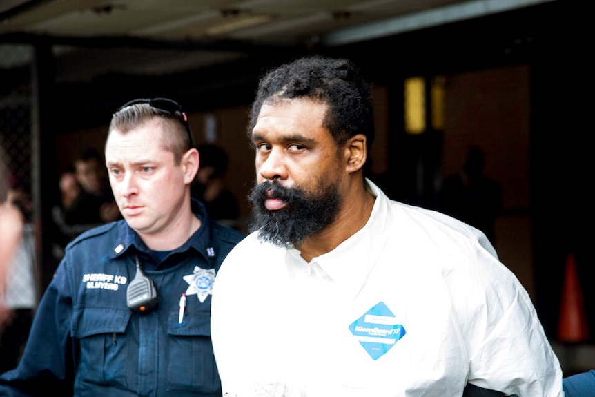 A man is pictured in what appears to be a white jumpsuit as he is escorted by police.