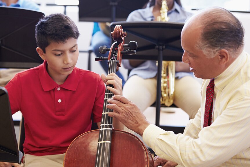A teacher helps a high school boy learn how to play a cello in a school orchestra.