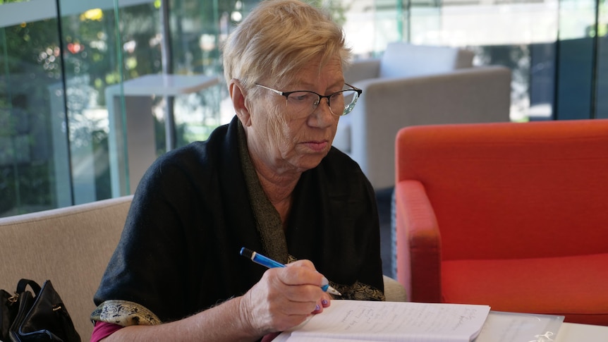 Woman with glasses sitting at a desk in a bright room holds a pen whilst looking down at her page.