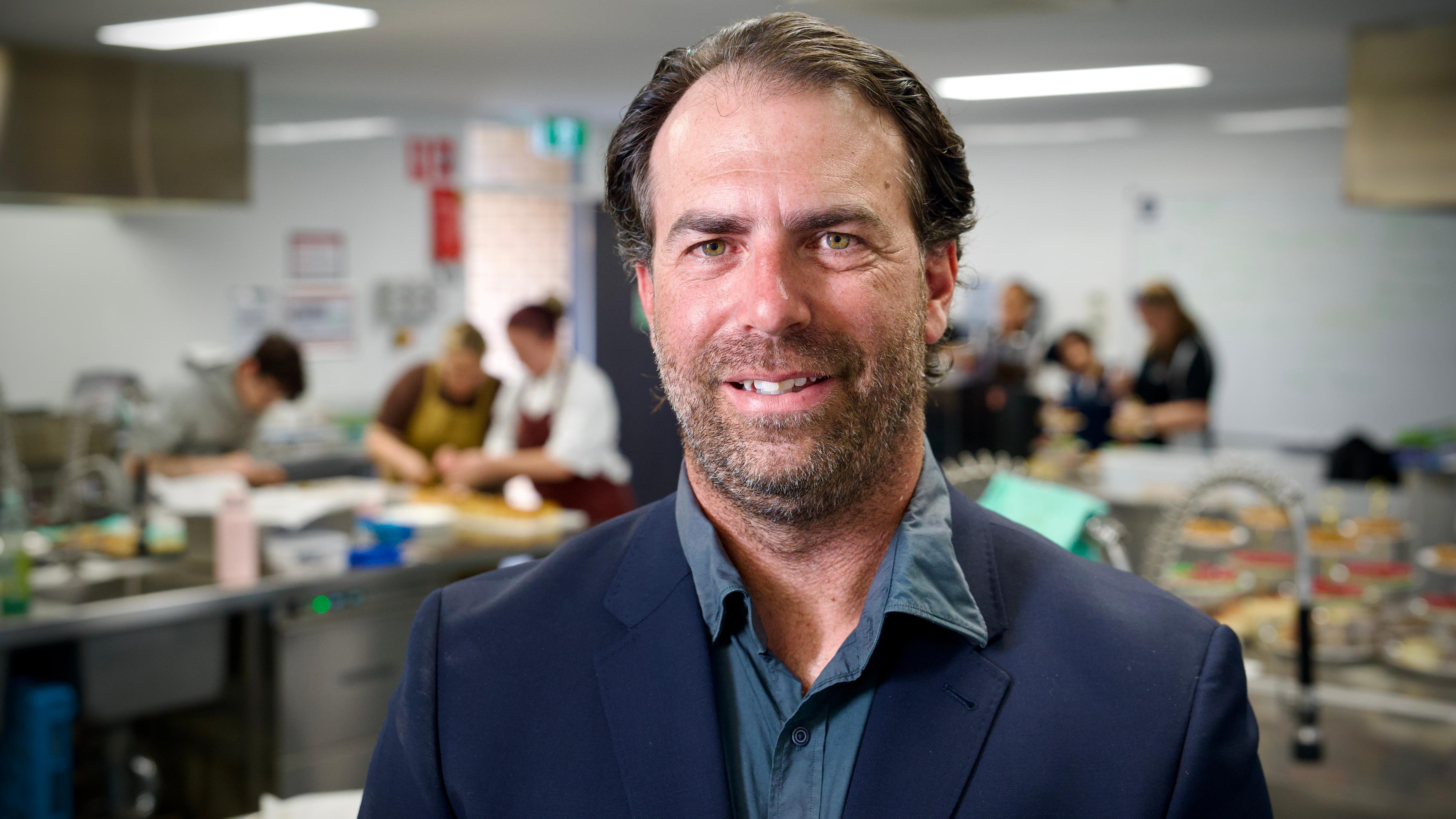 A man stands smiling at the camera in a commercial kitchen with chefs working behind him