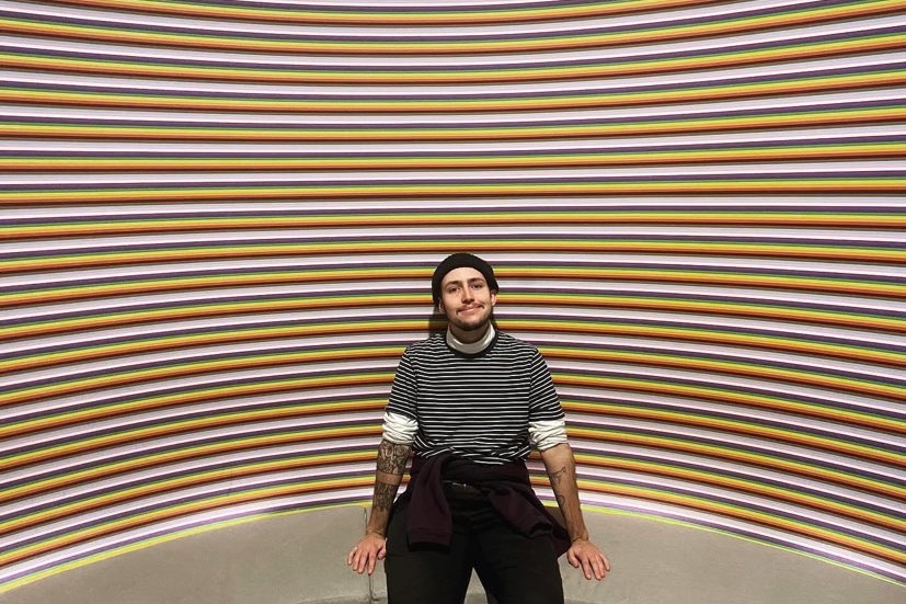 A male in his mid 20s in a stripy jumper and black beanie sits in front of a rainbow striped-wall and smiles.