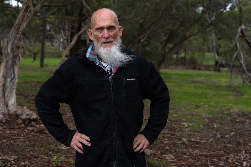 A man with a long grey beard wears a solemn expression while standing in a park