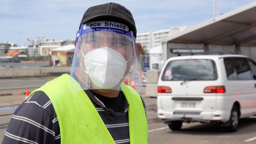 A man stands at a drive-through COVID-19 testing site wearing a yellow hi-vis vest, face shield and mask and hat.