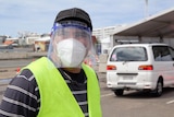 A man stands at a drive-through COVID-19 testing site wearing a yellow hi-vis vest, face shield and mask and hat.