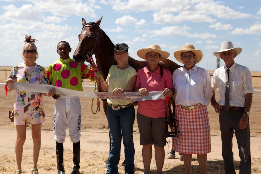 A group of people stand holding a long ribbon, in front of a racehorse on a dirt track
